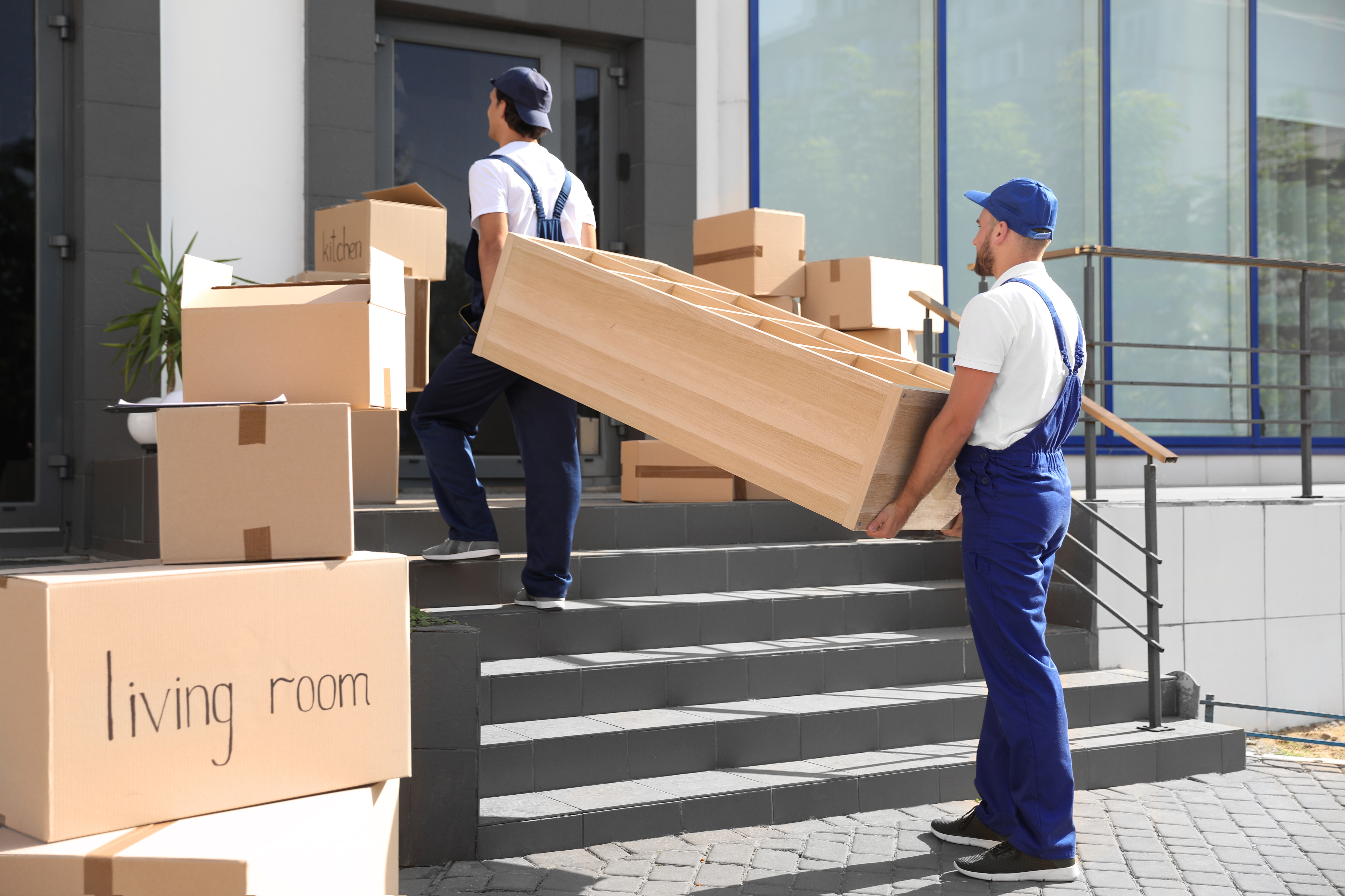 Two movers in blue uniforms carry a wooden dresser up steps. Several labeled boxes, including "living room" and "kitchen," are nearby.