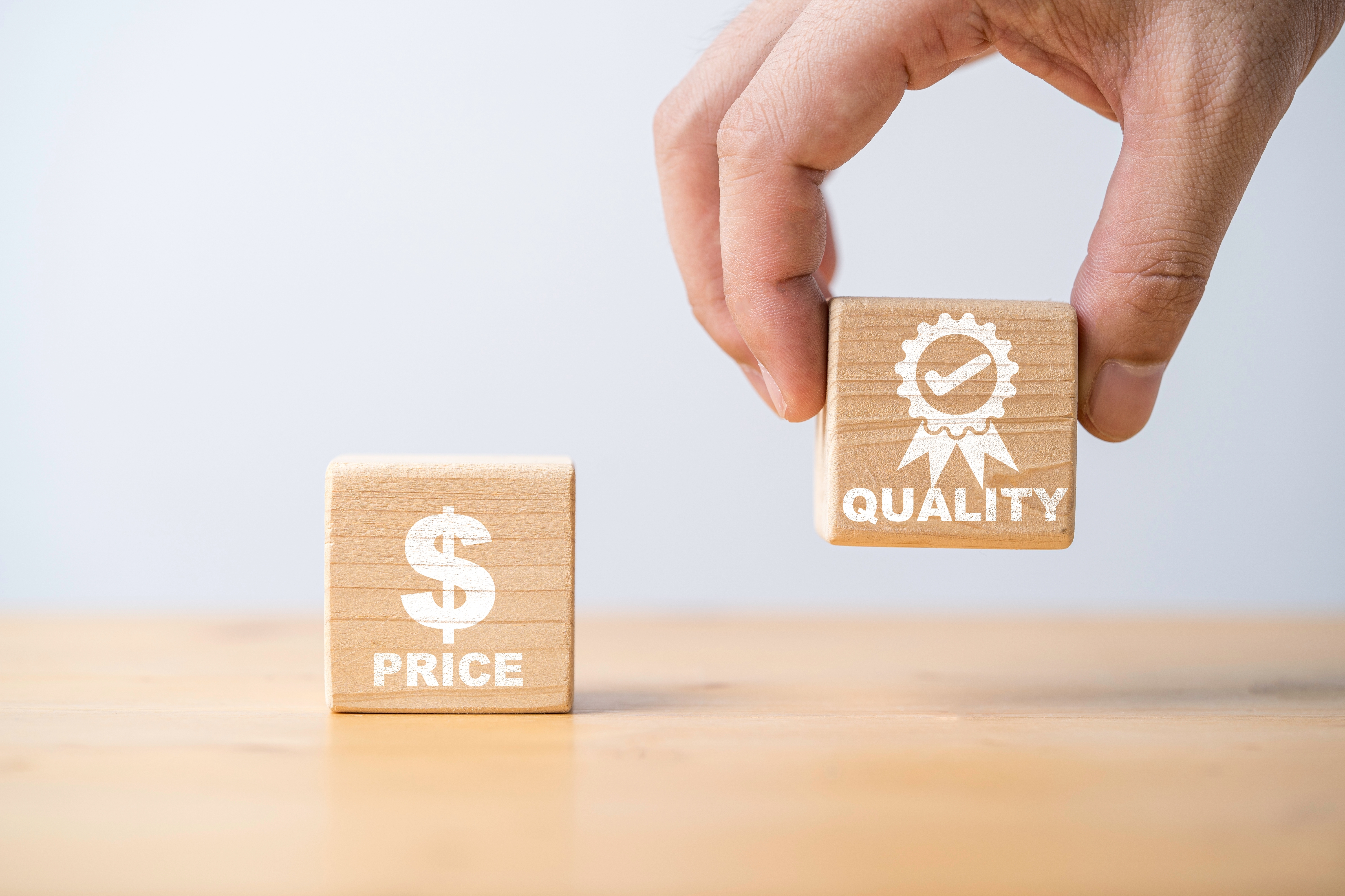 Hand holds a wooden block labeled "QUALITY" with a checkmark. Another block labeled "$ PRICE" sits on the table. Light background.