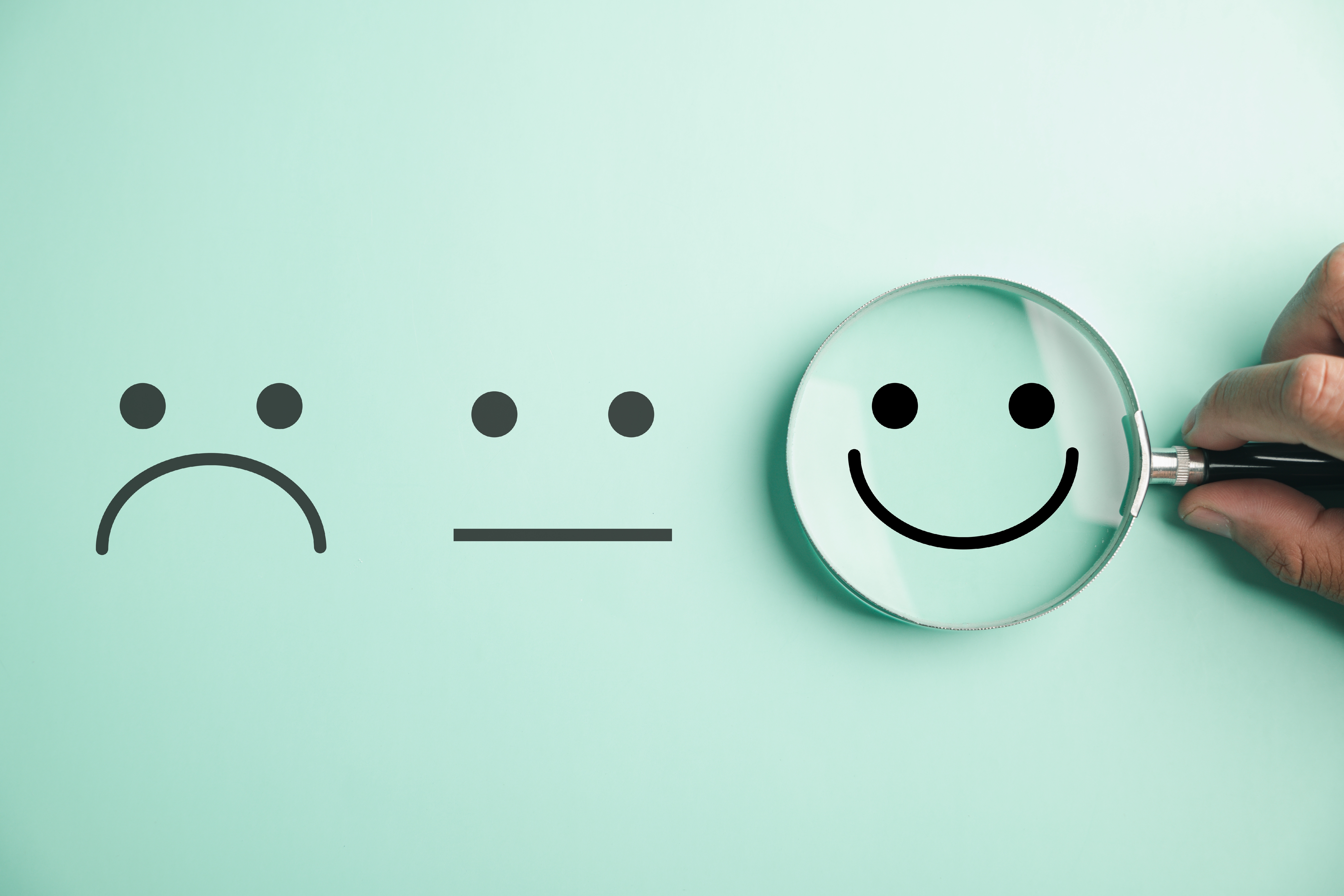 Sad, neutral, and happy faces on mint green background. A hand holds a magnifying glass, enlarging the happy face, suggesting positivity.