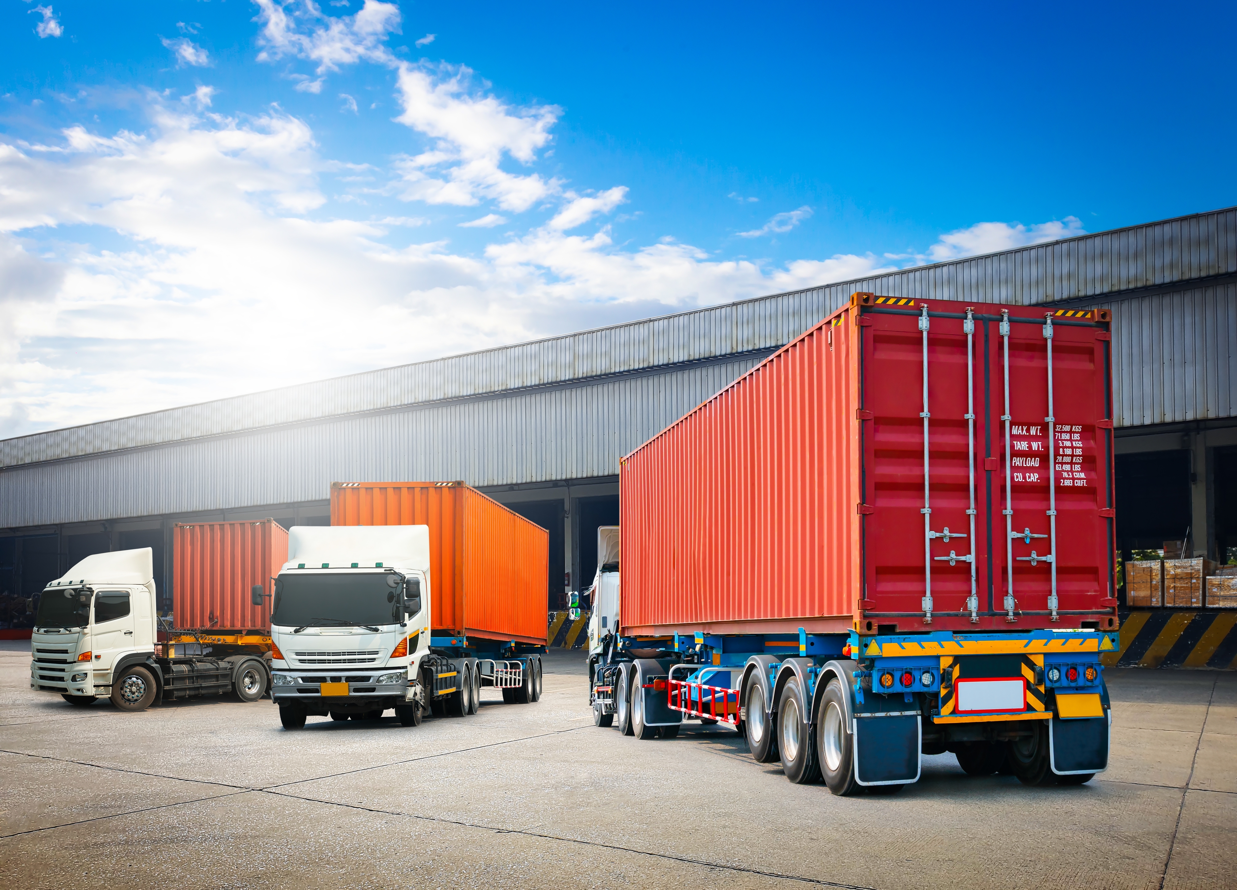 Three trucks with orange shipping containers parked near a warehouse under a bright blue sky. Text on containers is partially visible.