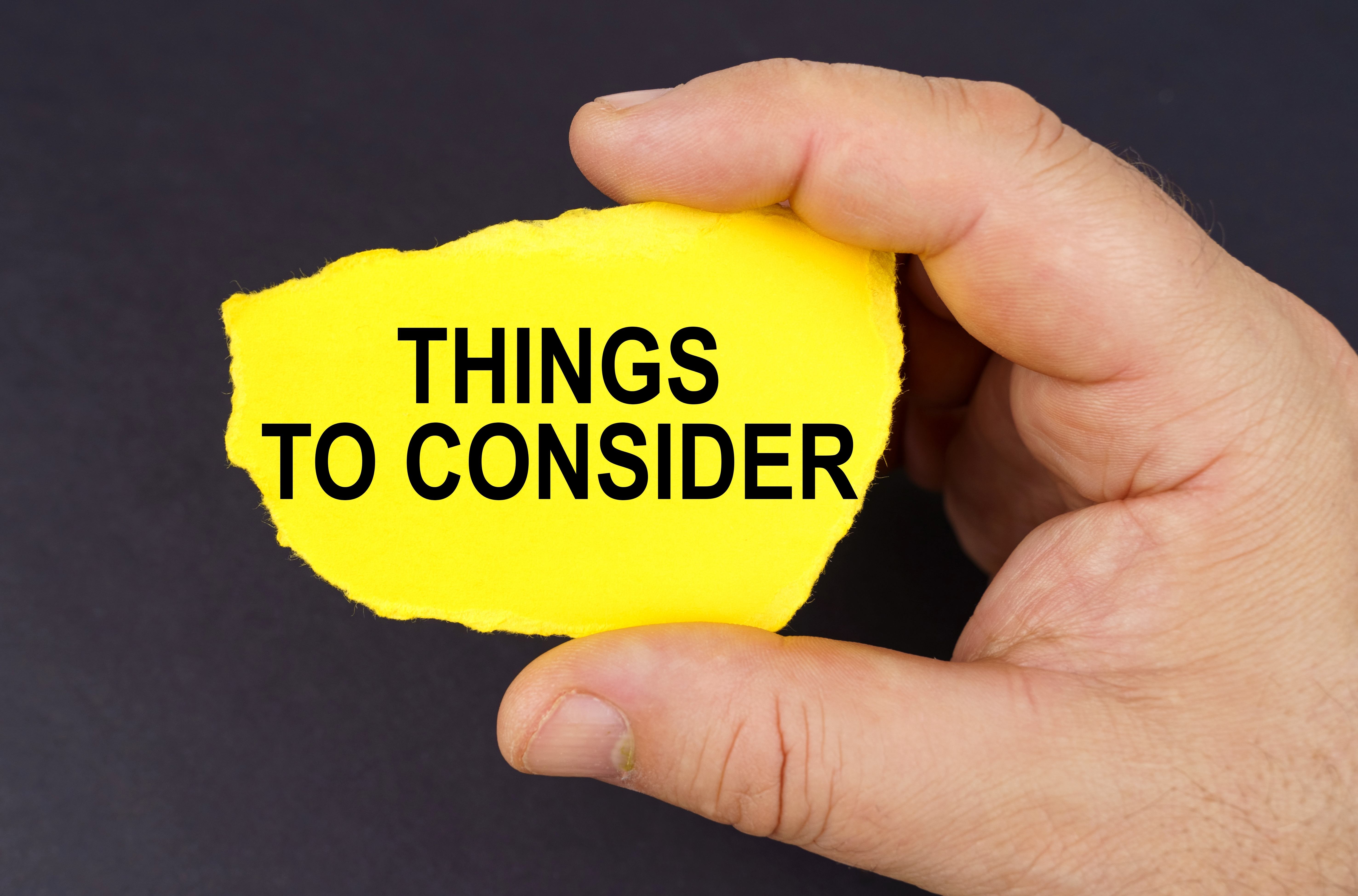 Hand holds yellow paper with "THINGS TO CONSIDER" text on dark background, conveying decision-making focus.