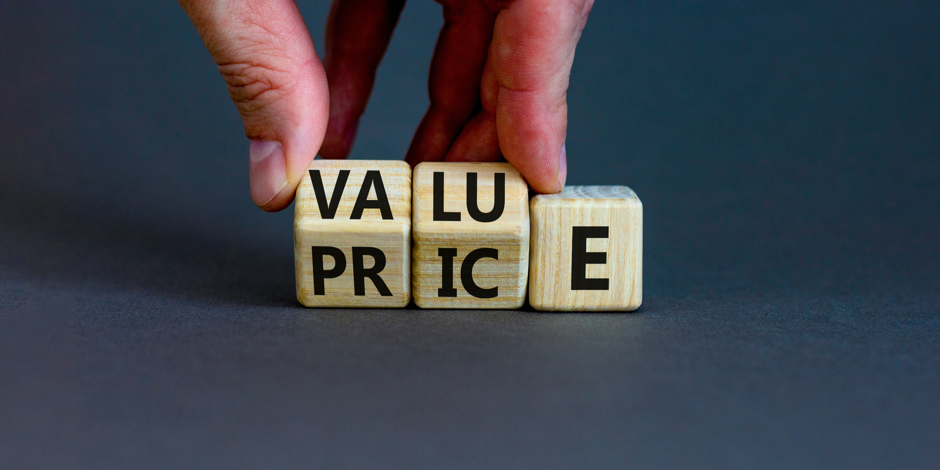 Hand rotates wooden blocks spelling PRICE to VALUE on a dark blue background, suggesting a change in focus or perspective.