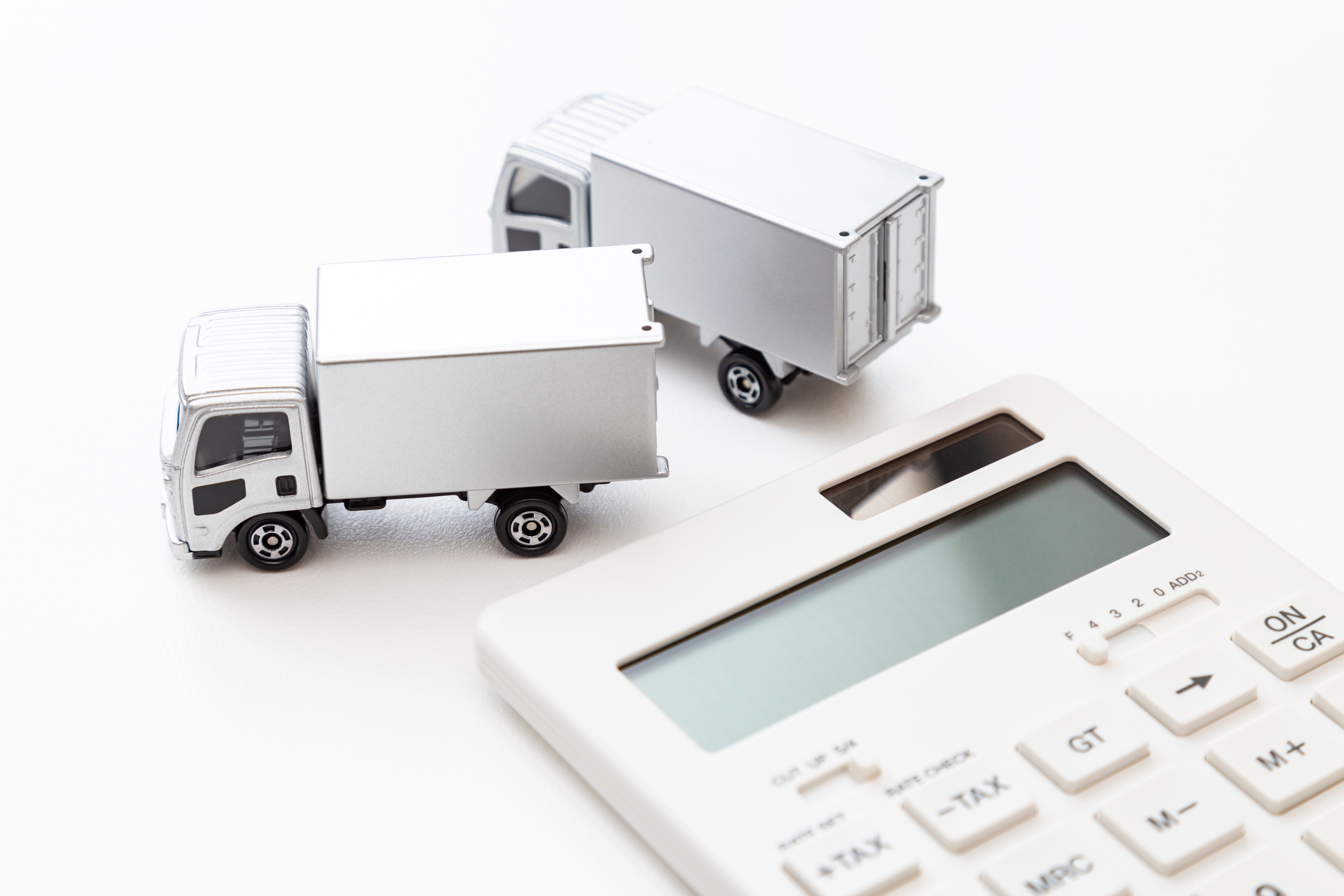 Two silver toy trucks and a white calculator on a bright surface. The calculator displays various buttons and text, creating a minimalistic scene.